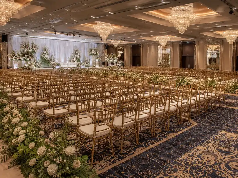Rows of gold Chiavari chairs in a hotel ballroom ceremony setup with chandelier lighting and floral aisle